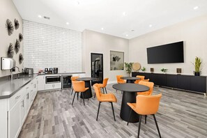 Eine moderne Küche mit weißen Schränken, einer schwarzen Arbeitsplatte und einer Spritzschutzwand. Zwei runde Tische mit orangefarbenen Stühlen stehen dort. Ein großer Flachbildfernseher ist an der Wand befestigt.