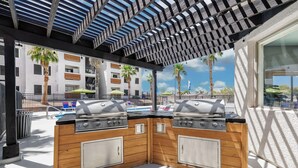 Uma área de churrasco externa com duas churrasqueiras a gás, armários de madeira e uma pérgola.