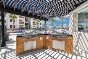 Ein Grillbereich im Freien mit zwei Gasgrills, Holzsäcken und einer Pergola darüber.