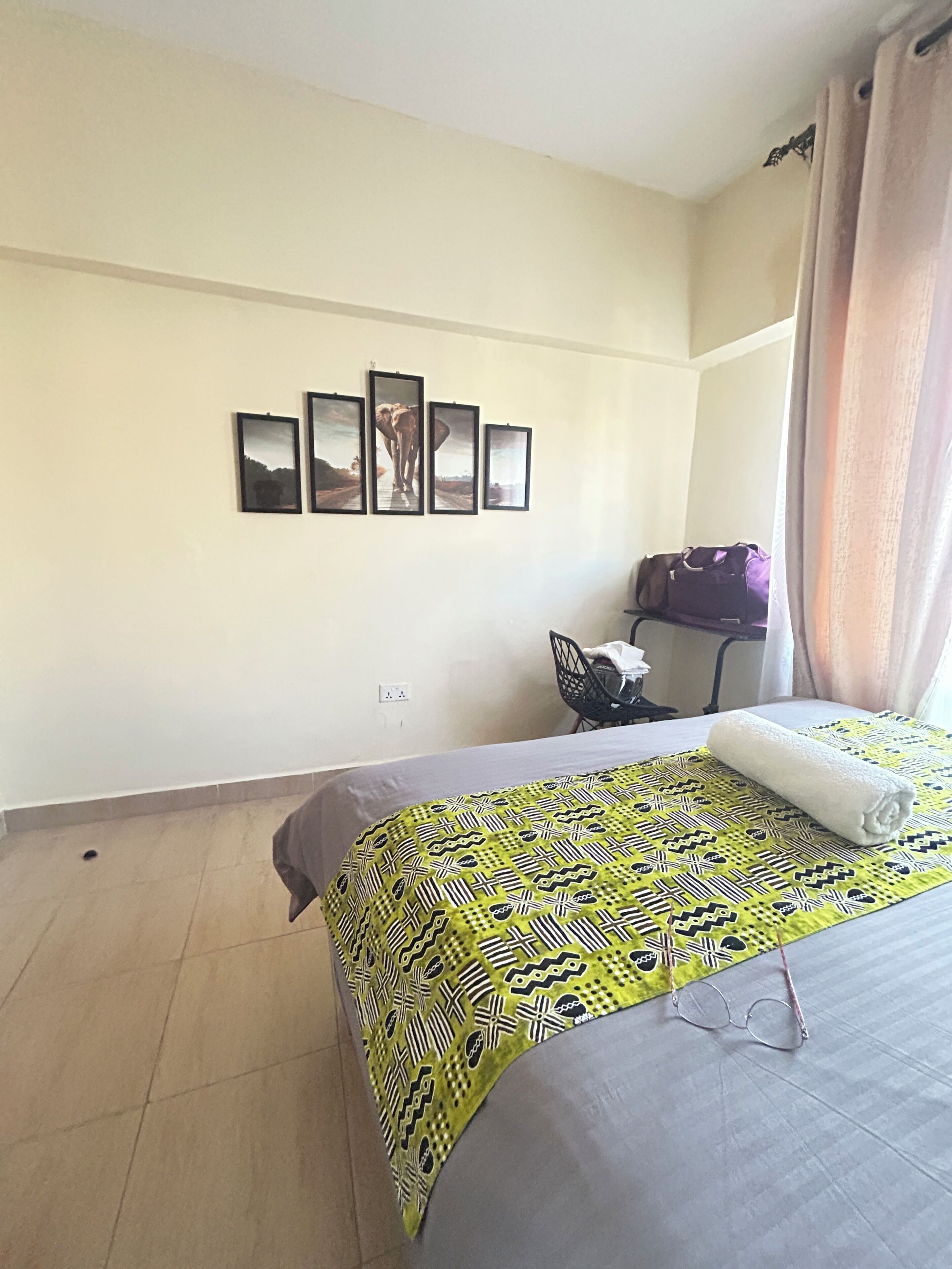 A hotel room with a bed, a chair, a purple bag, and four framed pictures on the wall.