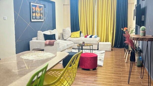 A modern living room with a sofa, ottoman, dining table, and chairs. There is a TV mounted on the wall and a ceiling light fixture. - Regal Suites (Athi River)