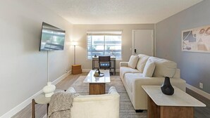 Ein modernes Wohnzimmer mit einer Couch, einem Couchtisch und einem Fernseher. Ein Fenster mit Jalousien, ein Schreibtisch und ein Stuhl sind ebenfalls vorhanden.