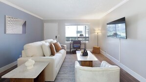 Ein modernes Wohnzimmer mit Sofa, Couchtisch und Sesseln. Es gibt einen Schreibtisch mit Stuhl, eine Lampe und einen an der Wand befestigten Fernseher.