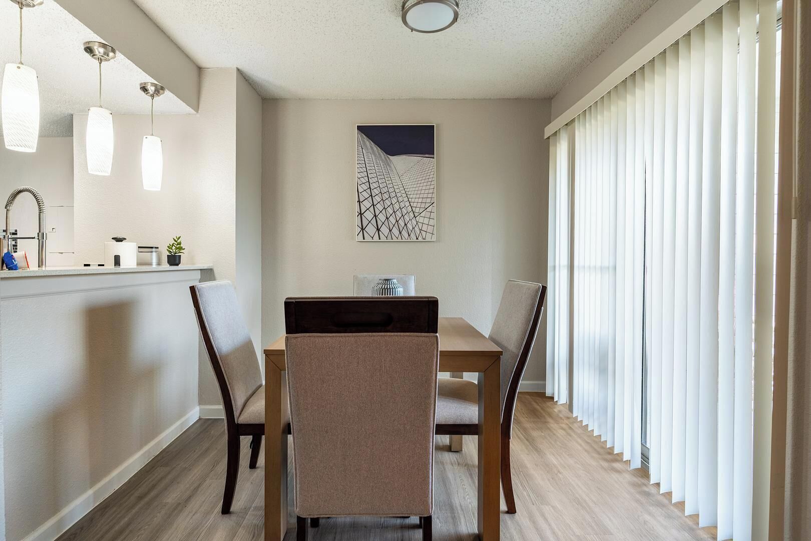 A dining area with a wooden table, four chairs, a kitchen counter with a sink and cabinets, and a large window with blinds.
