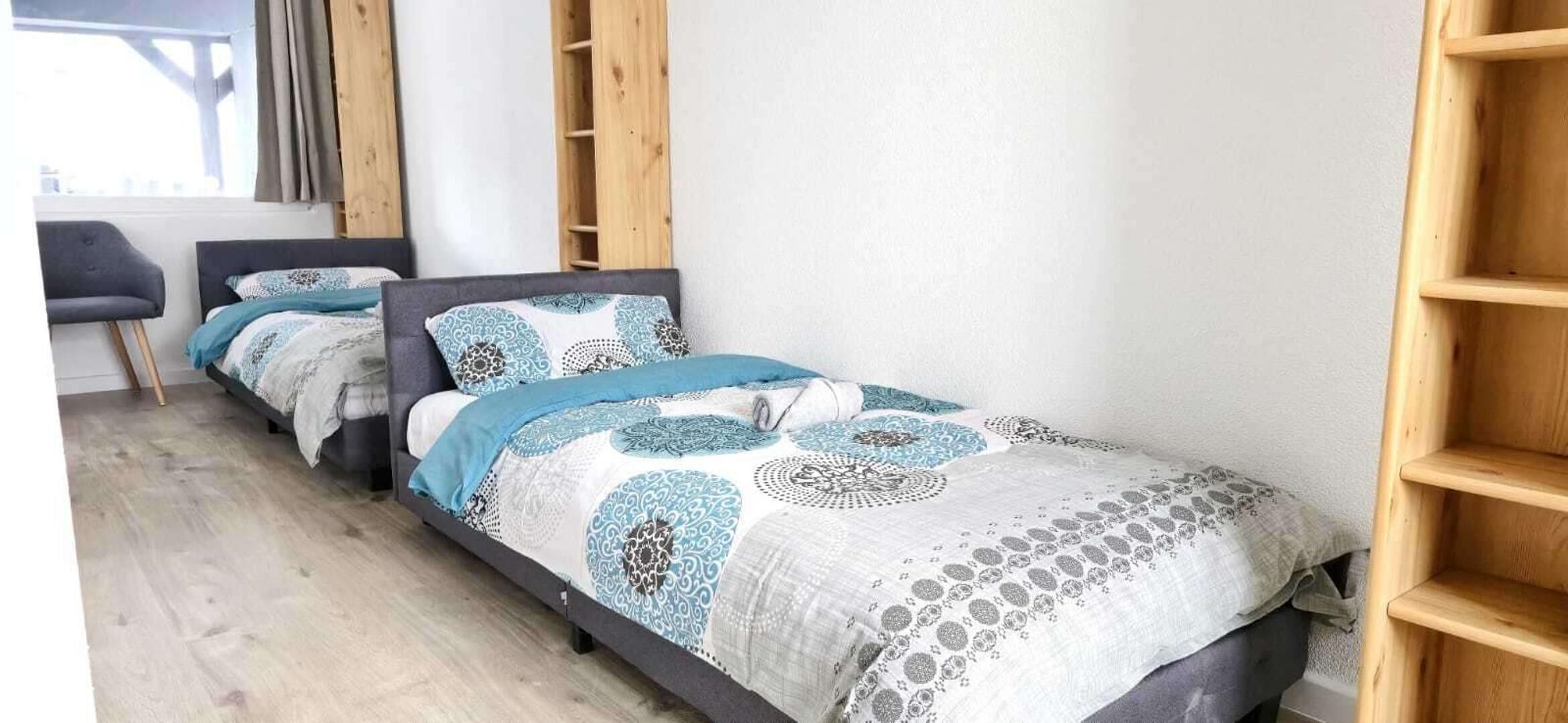 A small, modern room with two single beds, a grey chair, and a wooden shelf unit.