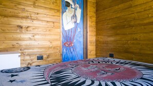 A wooden cabin interior with a bed featuring a large, colorful tapestry.