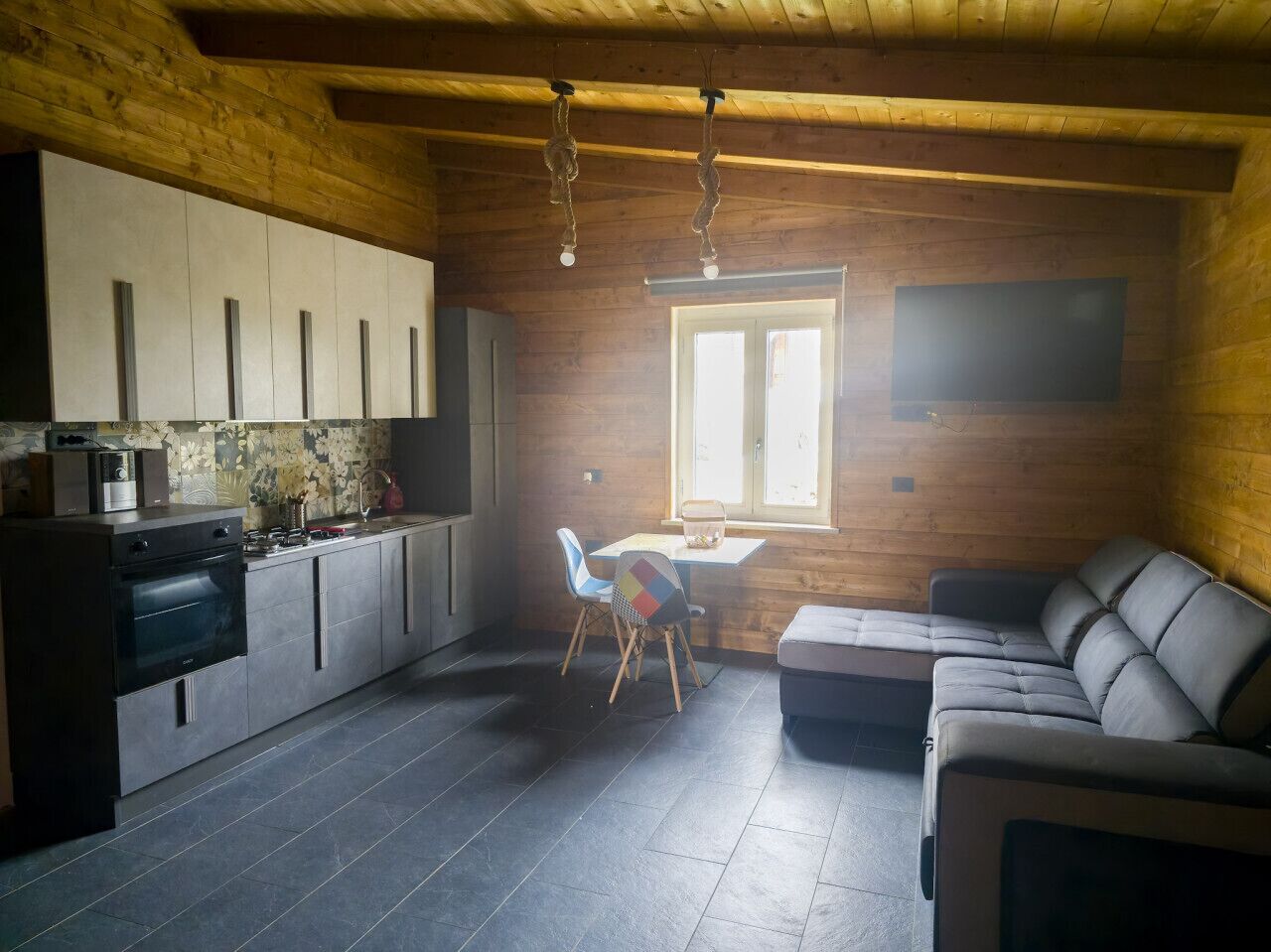 Une cuisine moderne avec des carreaux foncés, des meubles en bois et un four encastré. On y trouve un coin repas avec une table et des chaises, ainsi qu'un canapé dans l'espace salon.
