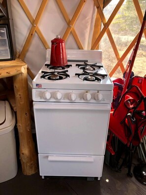 A white stove with four burners and a red kettle on top, located in a tent-like structure with wooden beams.