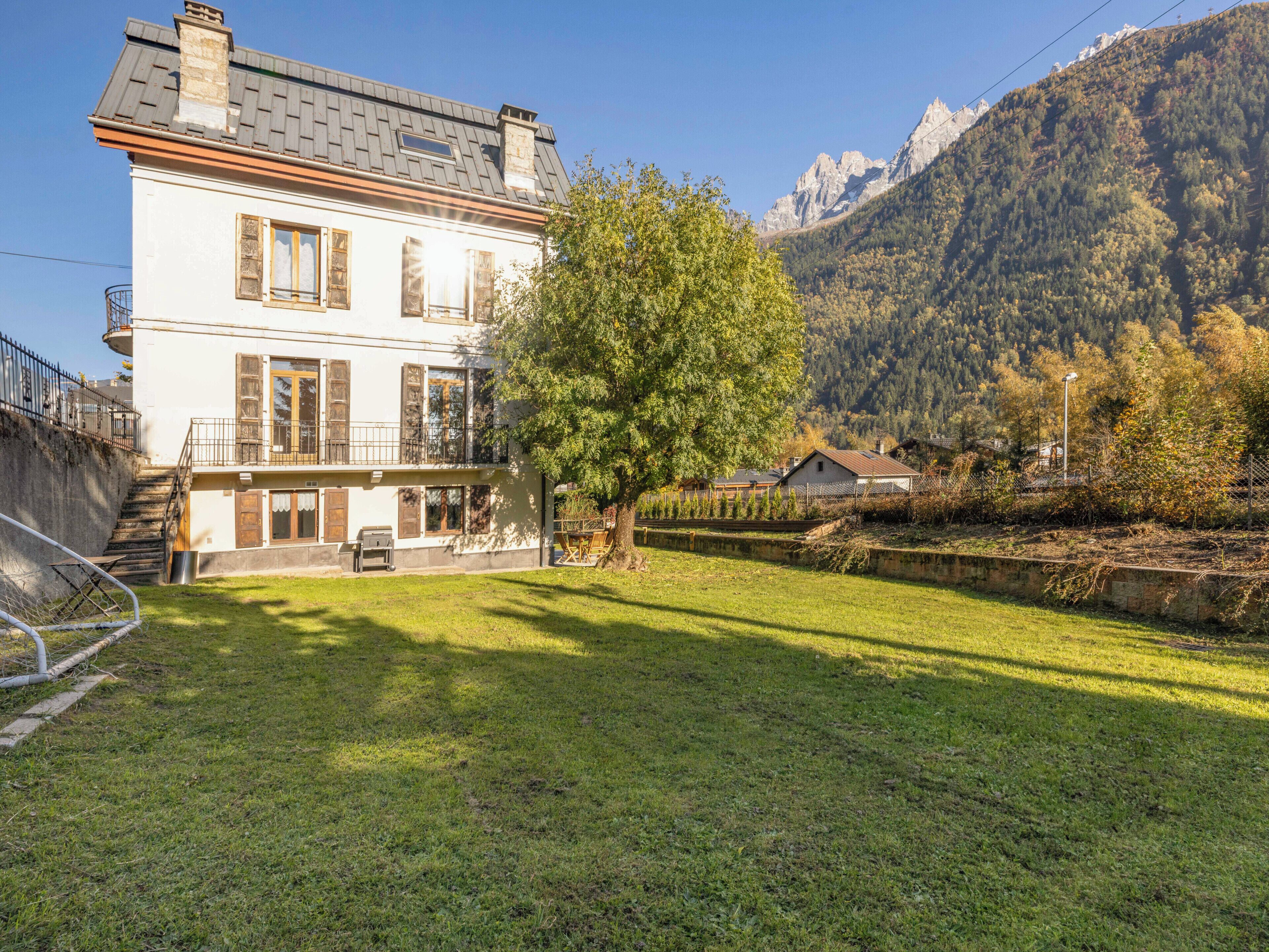 Une maison à deux étages avec un balcon, un arbre dans la cour et des montagnes en arrière-plan.