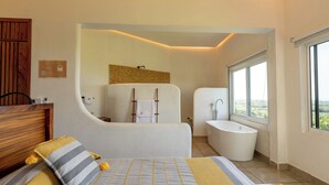 A modern bedroom with a large bed, a wooden headboard, a bedside table, a nightstand, a large window, and a bathroom with a bathtub and sink. - Casa Cumbre PXM (Santa María Colotepec)