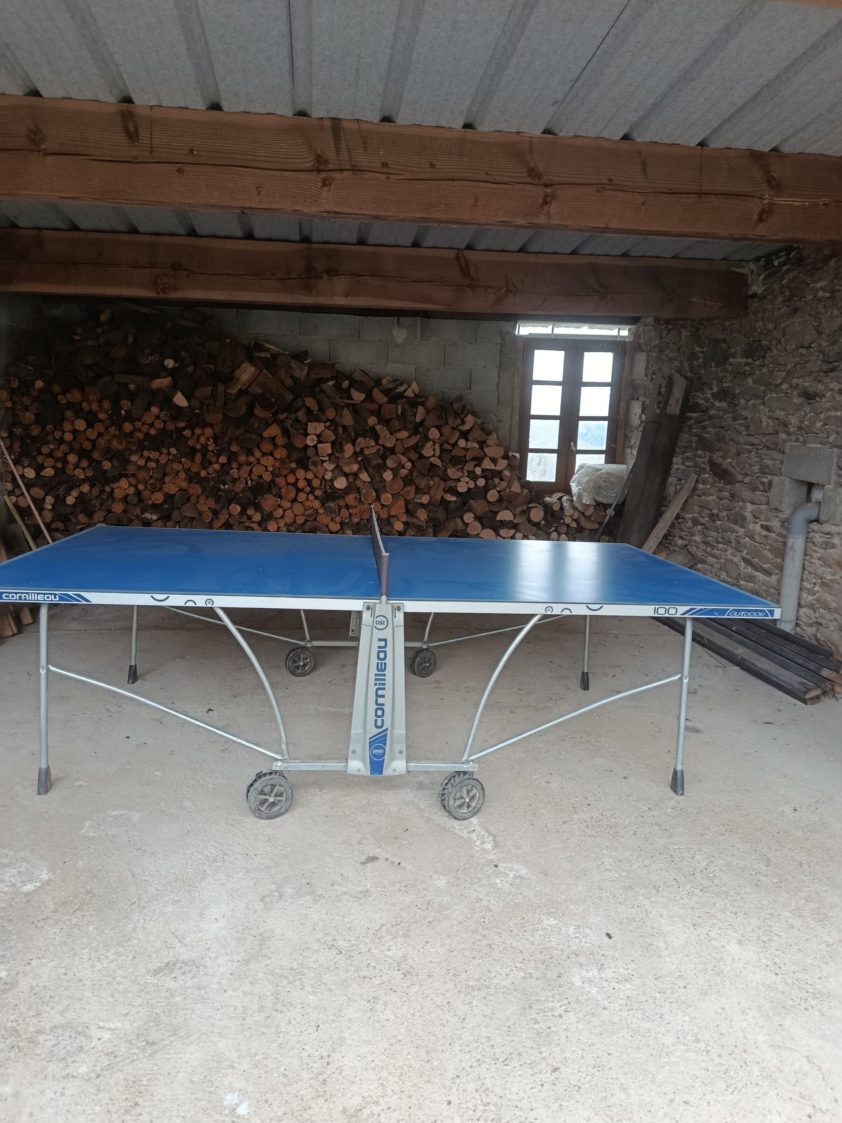 Une table de ping-pong dans une pièce rustique, avec un mur en pierre et une pile de bois de chauffage.