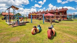 A playground with colorful equipment and tires on the grass, a building with multiple red roofs, and a clear blue sky. - Residencial Encantos Linha Bonita (Gramado)