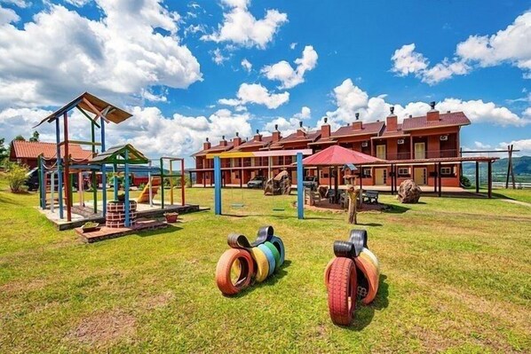 Um parquinho com equipamentos coloridos e pneus na grama, um edifício com vários telhados vermelhos e um céu azul claro.