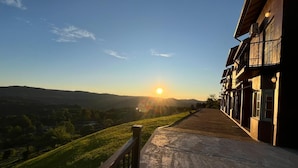 A building with a paved pathway leading to a grassy area with a view of the sun setting over a hilly landscape. - Residencial Encantos Linha Bonita (Gramado)