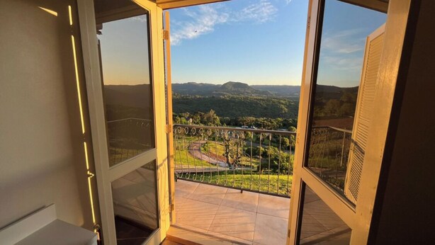 A room with a large glass door leading to a balcony with a view of a landscape and a bridge. - Residencial Encantos Linha Bonita (Gramado)