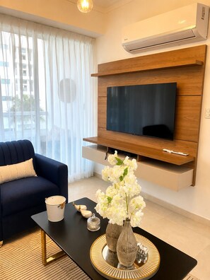 Ein modernes Wohnzimmer mit einem flachbildfernseher an einer Holzwand, einem dunklen Sofa, einem Couchtisch mit Dekoration und transparenten Vorhängen.