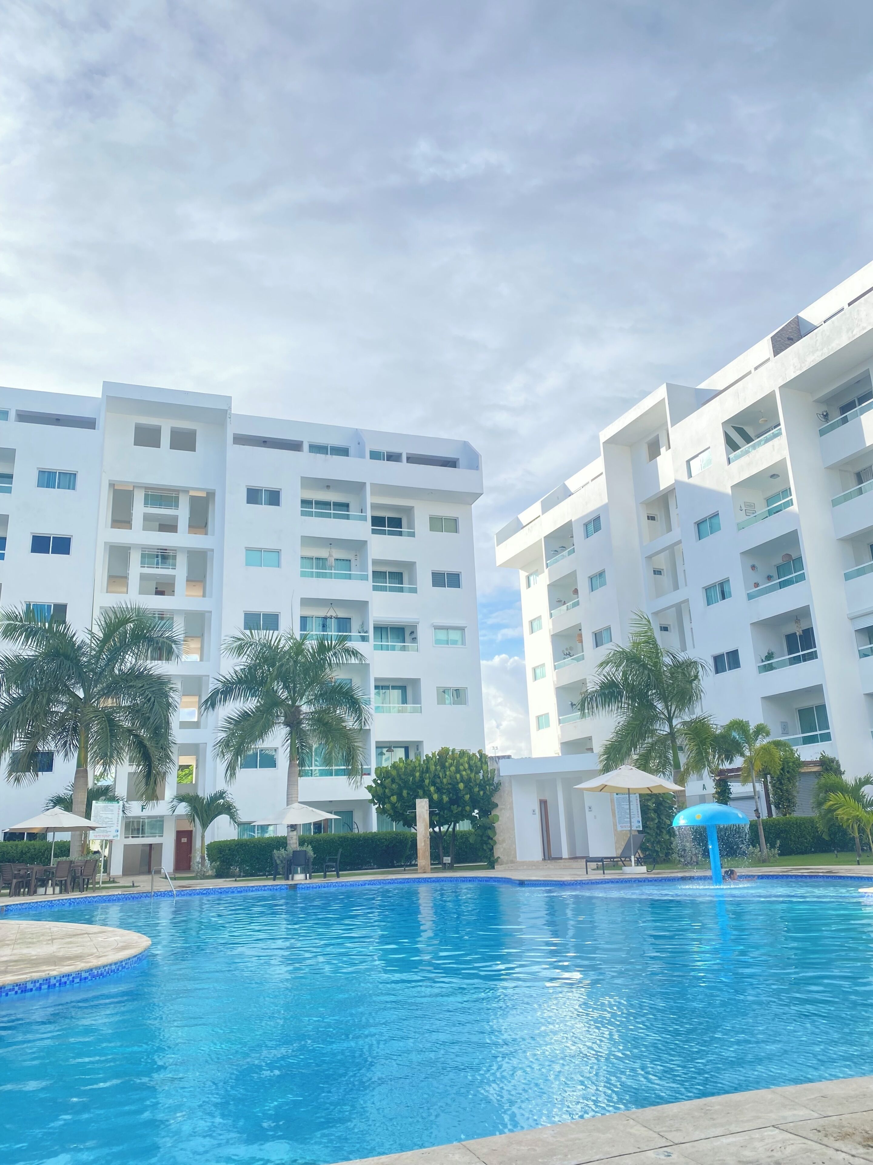 Une piscine bordée de bleu, entourée de palmiers et d’un bâtiment blanc doté de nombreux balcons.