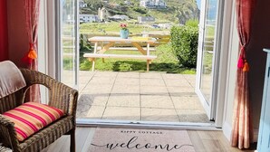 A wicker chair with a striped cushion and a welcome mat in front of a sliding glass door leading to a patio with a picnic table and a view of houses on a hillside. - Kippy Cottage (Truro)