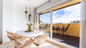 Een balkon met een zithoek, bestaande uit wickerstoelen en een ronde tafel, met een uitzicht op gebouwen op de achtergrond.