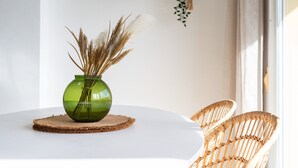 Een ronde tafel met een groene vaas met gedroogde planten, een geweven placemat en een rieten stoel.
