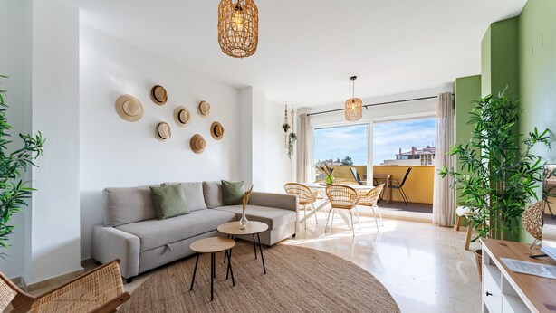 Een moderne woonkamer met een bank, ronde tafels, rieten stoelen en een groot raam met uitzicht.