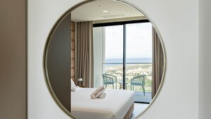Habitación de hotel con un espejo grande, una cama con cubrecama blanco impecable, dos sillas verdes y un balcón con vistas a edificios.