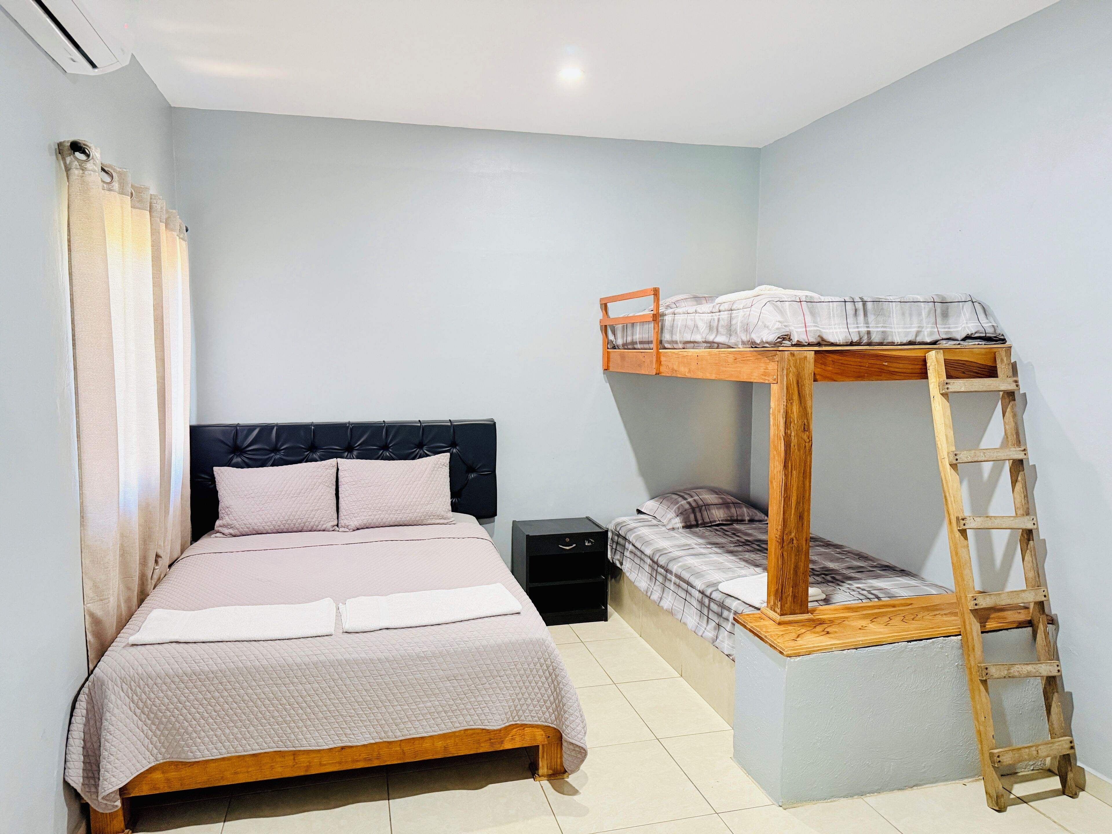 Une pièce avec un lit superposé, un lit en bas, une échelle en bois, une petite armoire noire et un rideau.