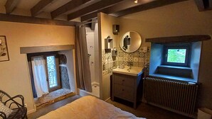 Un dormitorio con cama, una silla, un ventanal pequeño, un radiador y un baño con lavabo y espejo.