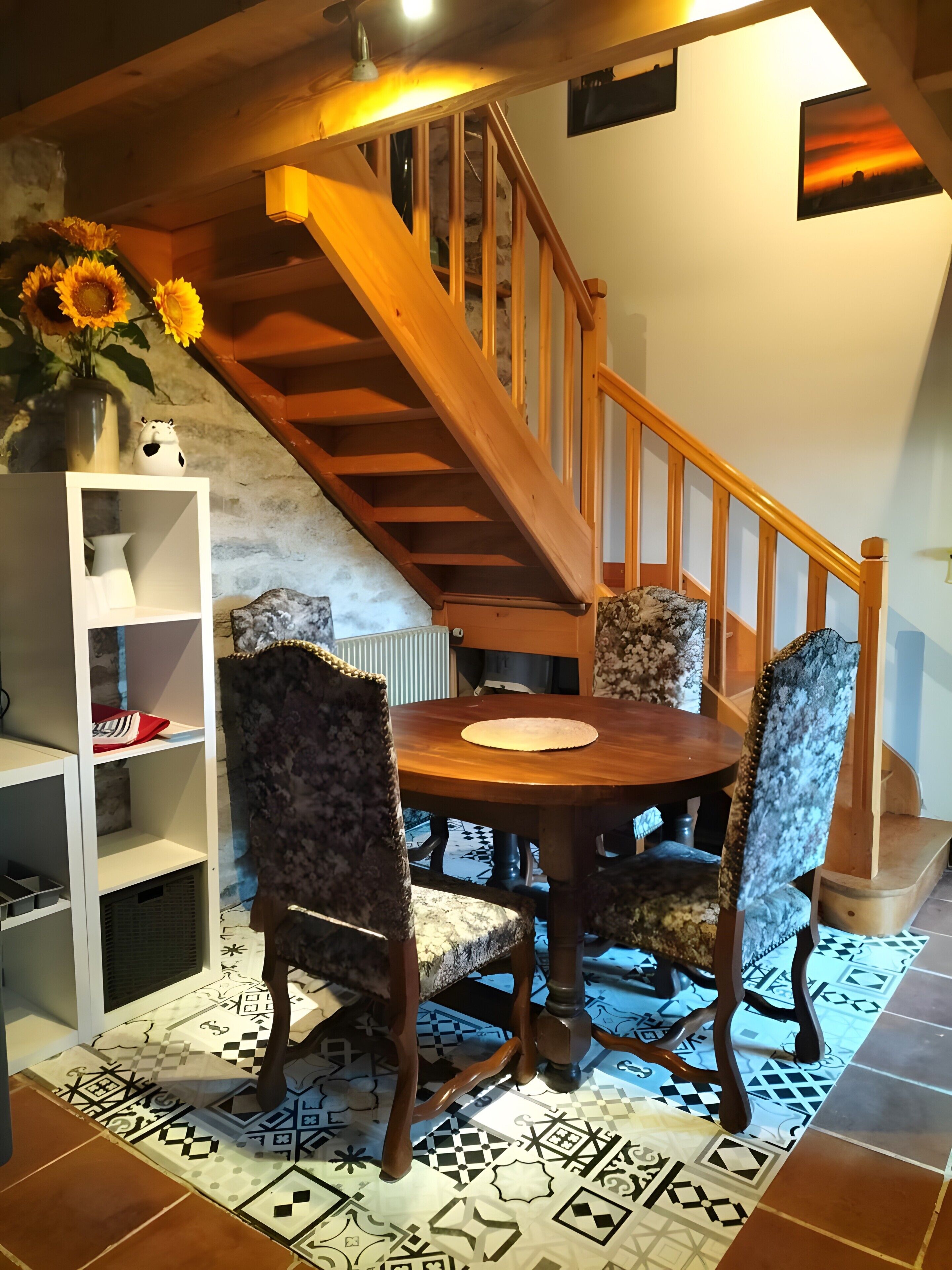 Un espace repas comprenant une table en bois, des chaises à motifs, une étagère blanche et un escalier.