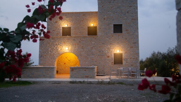 A stone building with an arched entrance, a small outdoor seating area with chairs, and a stone wall. - Anaroussa Diros Villa (East Mani)