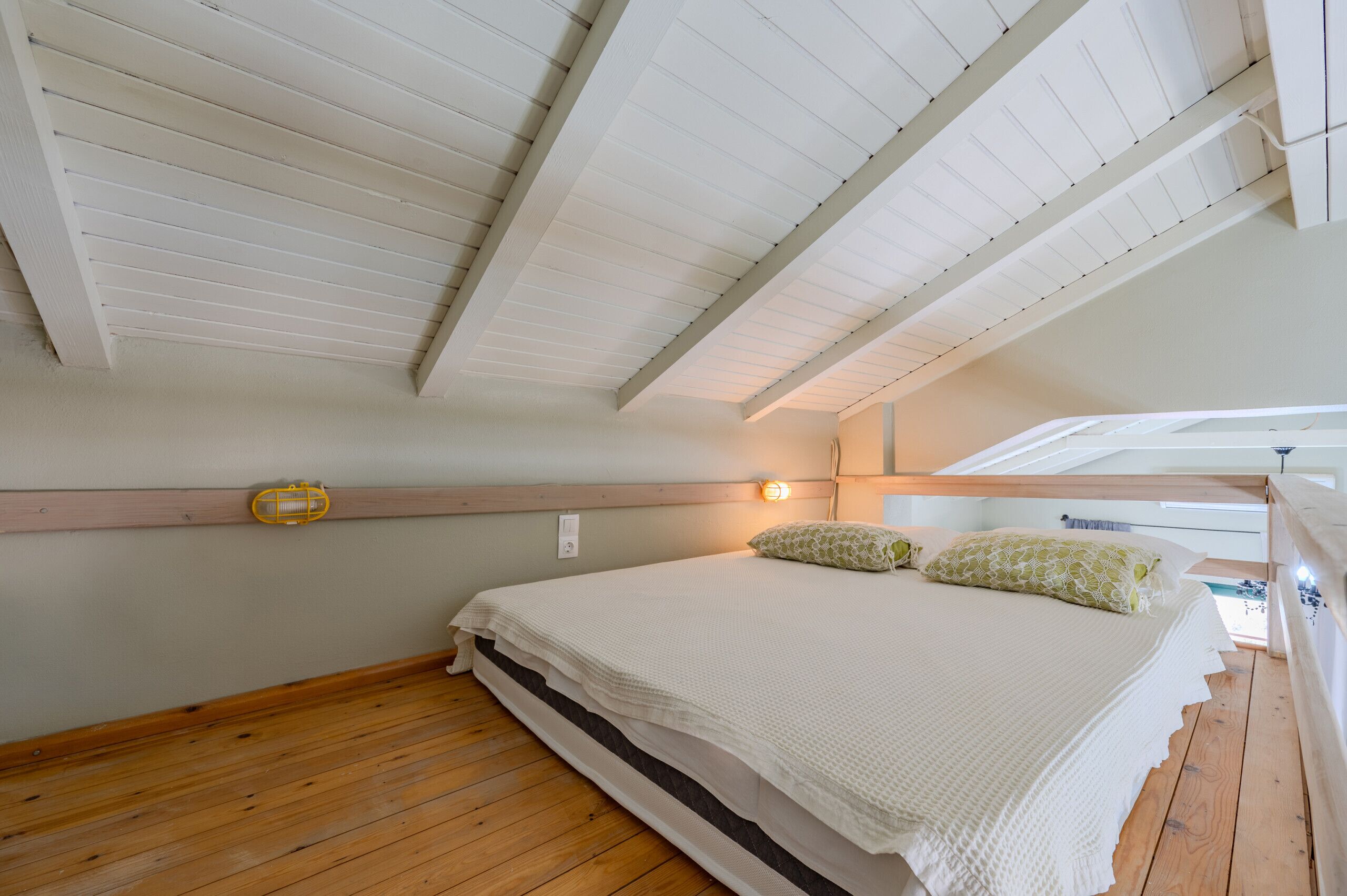 Una camera da letto con soffitto spiovente, un letto con copripiumino bianco, due cuscini e pavimento in legno.