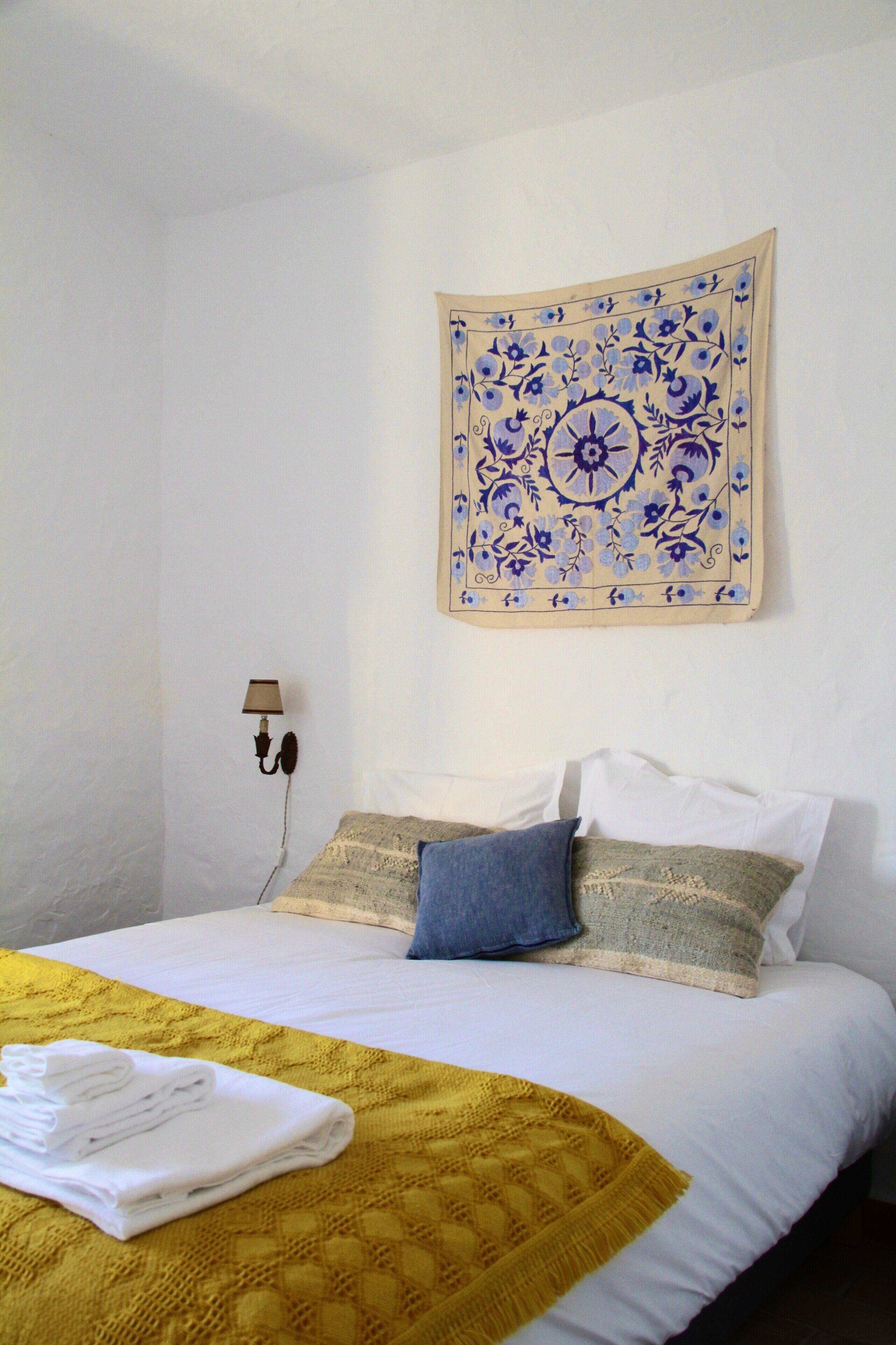 A neatly made bed with a yellow blanket, white pillows, and a blue pillow, a wall tapestry, and a wall-mounted lamp.