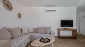 A modern living room with a beige sectional sofa, a wooden coffee table with decorative items, a flat-screen TV on a wooden stand, and an air conditioning unit mounted on the wall. - Mythos 4 (Agios Vasileios)