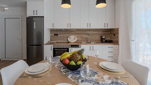 A modern kitchen with white cabinets, a stainless steel refrigerator, wooden accents, and a fruit bowl on the dining table. - Mythos 4 (Agios Vasileios)