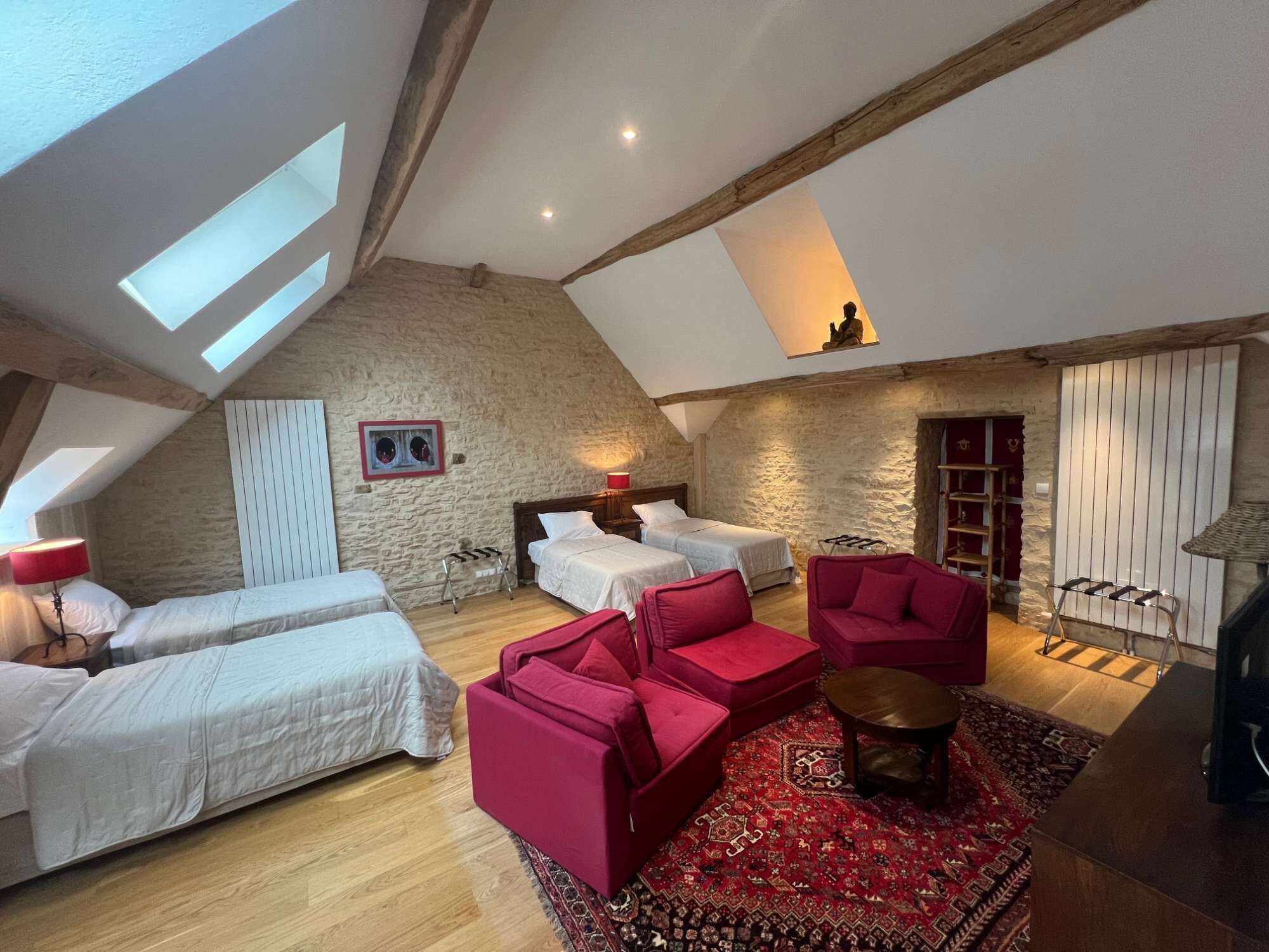 A spacious loft bedroom with a high ceiling, exposed wooden beams, and skylights. It features two beds, a red sofa, and a patterned rug.