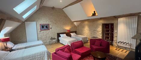 A spacious loft bedroom with a high ceiling, exposed wooden beams, and skylights. It features two beds, a red sofa, and a patterned rug.