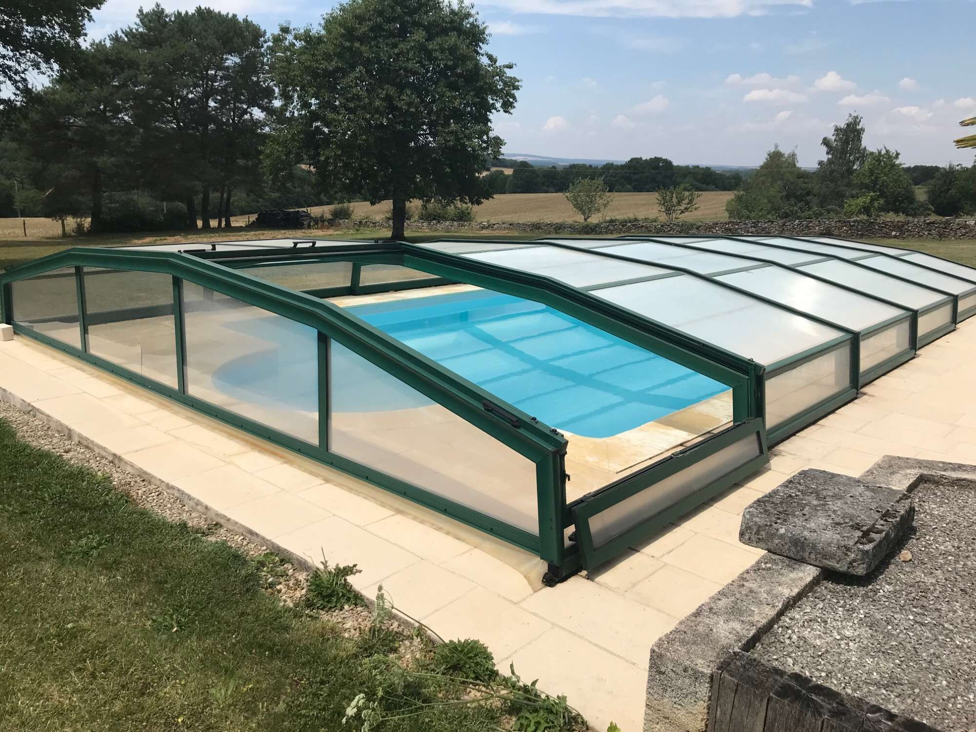 Une piscine dotée d’une enceinte en verre et d’une structure verte, entourée d’une zone pavée et d’arbres.