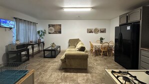 Una sala de estar con un sofá, una mesa de comedor con sillas, una zona de cocina con un refrigerador y un televisor montado en la pared.