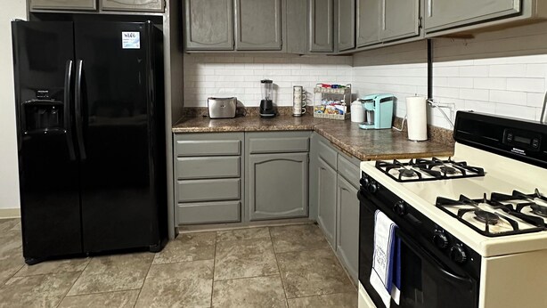 Una cocina con armarios grises, un refrigerador negro, una estufa y una encimera con varios electrodomésticos.