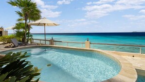 A pool with clear blue water, surrounded by a deck with lounge chairs and umbrellas, overlooking a turquoise sea and a clear sky. - Bellevue Penthouse 10 (Kralendijk)