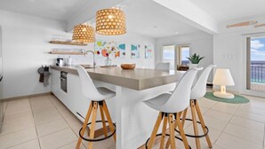 A modern kitchen with a central island, white bar stools, pendant lighting, and a view to the outside. - Bellevue Penthouse 10 (Kralendijk)