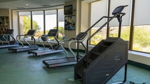 Uma academia com esteiras e uma máquina de escada, grandes janelas e piso verde.