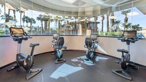 Un gimnasio con cuatro bicicletas estáticas, ventanas amplias y vistas a palmeras y edificios.