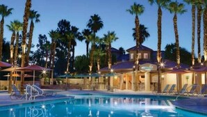Un área de piscina con sillones de descanso y sombrillas, rodeada de palmeras y un edificio de varios niveles.