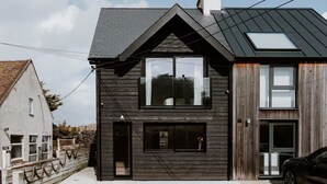 Ein modernes, zweistöckiges Haus mit dunkler Fassade, großen Fenstern und einem geparkten Auto davor.