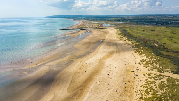Eine Vogelperspektive einer Küstenregion mit Sandstrand, blauem Himmel und sanften Hügeln im Hintergrund.