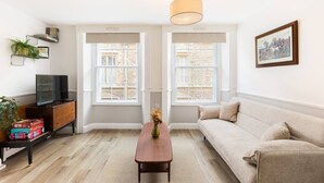 Un salon comprenant un canapé, une table basse, un téléviseur posé sur un meuble en bois, et deux fenêtres à cadres blancs.