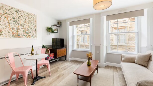 Un salon moderne avec un canapé blanc, une table basse en bois et un fauteuil rose. Un tableau est accroché au mur, il y a aussi un téléviseur et une fenêtre donnant sur des immeubles.