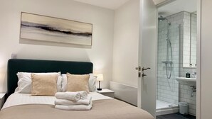 A modern bedroom with a large bed, a nightstand, a wall-mounted painting, and a bathroom with a shower and sink. - Converted Ancoats Mill - Walk to Victoria - Pass the Keys (Manchester)
