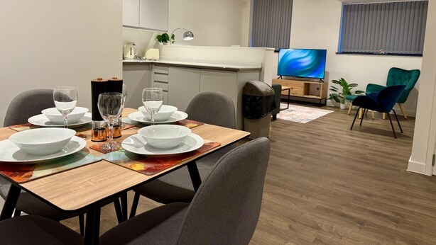 A modern dining area with a wooden table set for four, a kitchen in the background, a television, and a teal chair. - Converted Ancoats Mill - Walk to Victoria - Pass the Keys (Manchester)